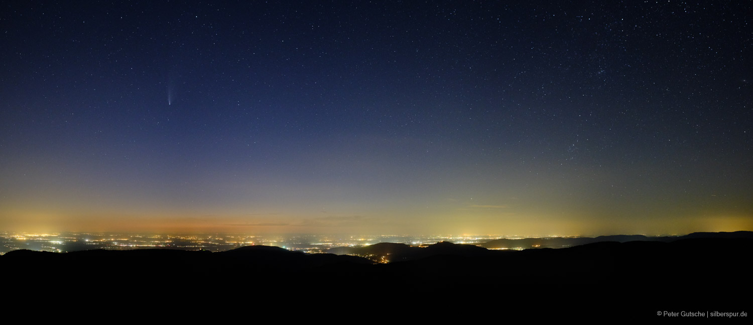 Night view from a high Black Forest mountain overlooking the Karlsruhe region. The entire horizon is illuminated by artificial lights. Many stars are visible in the sky, with the tail of Comet Neowise on the left.