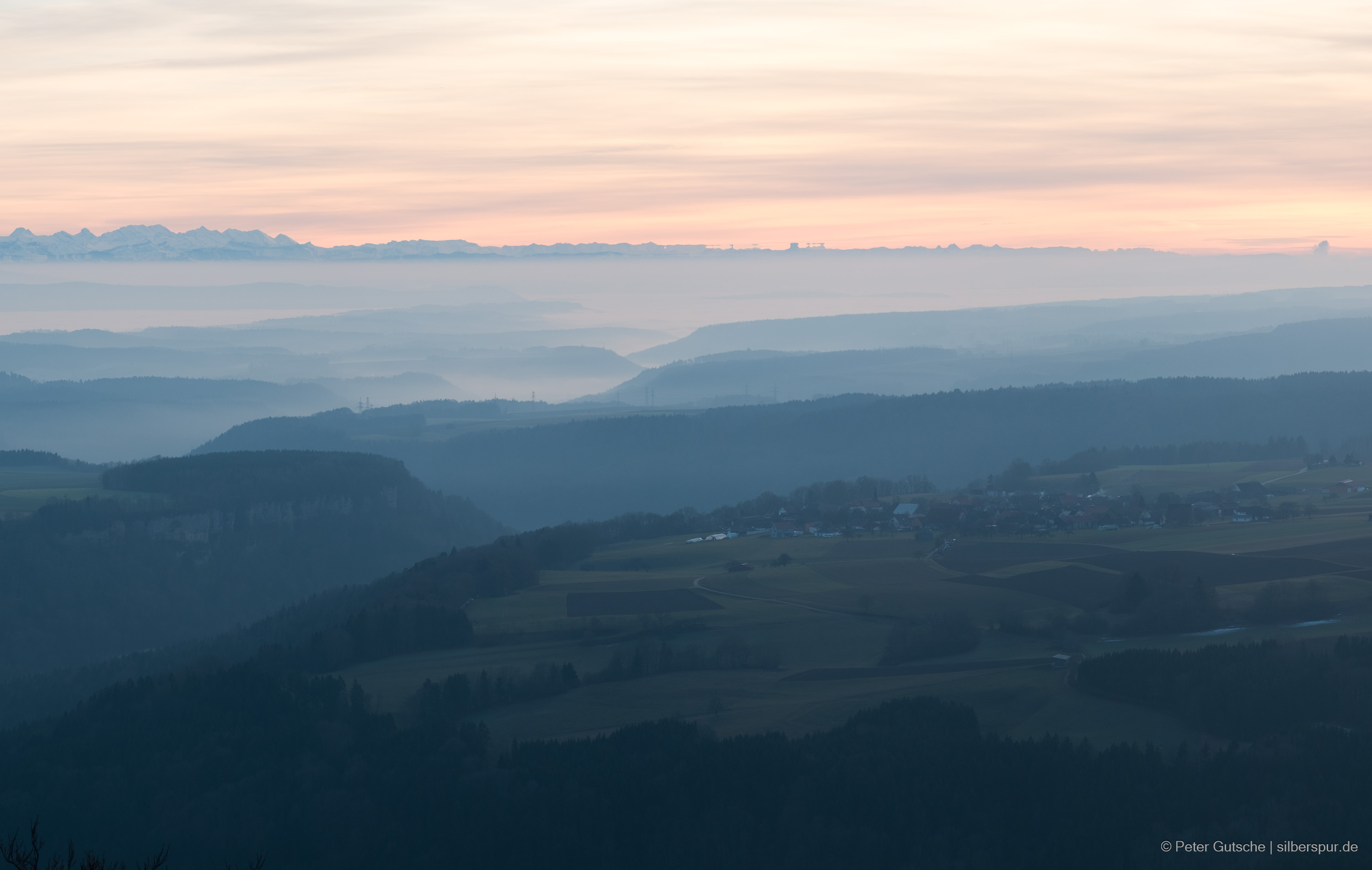 View from Buchberg in the Southern Black Forest towards the southwest against the afternoon sun. In the foreground, the lower hills of the Southern Black Forest are visible, with a village situated on the plateau. The sun is already so low that no direct light can reach any more parts of the landscape, therefore, the picture is in a blueish monochrome tone. Mist and fog lingers in the valleys below. The plateau and hilly landscape are deeply carved by the valleys of the Wutach River and its tributaries. In the background, the majestic panorama of the Alps can be seen.