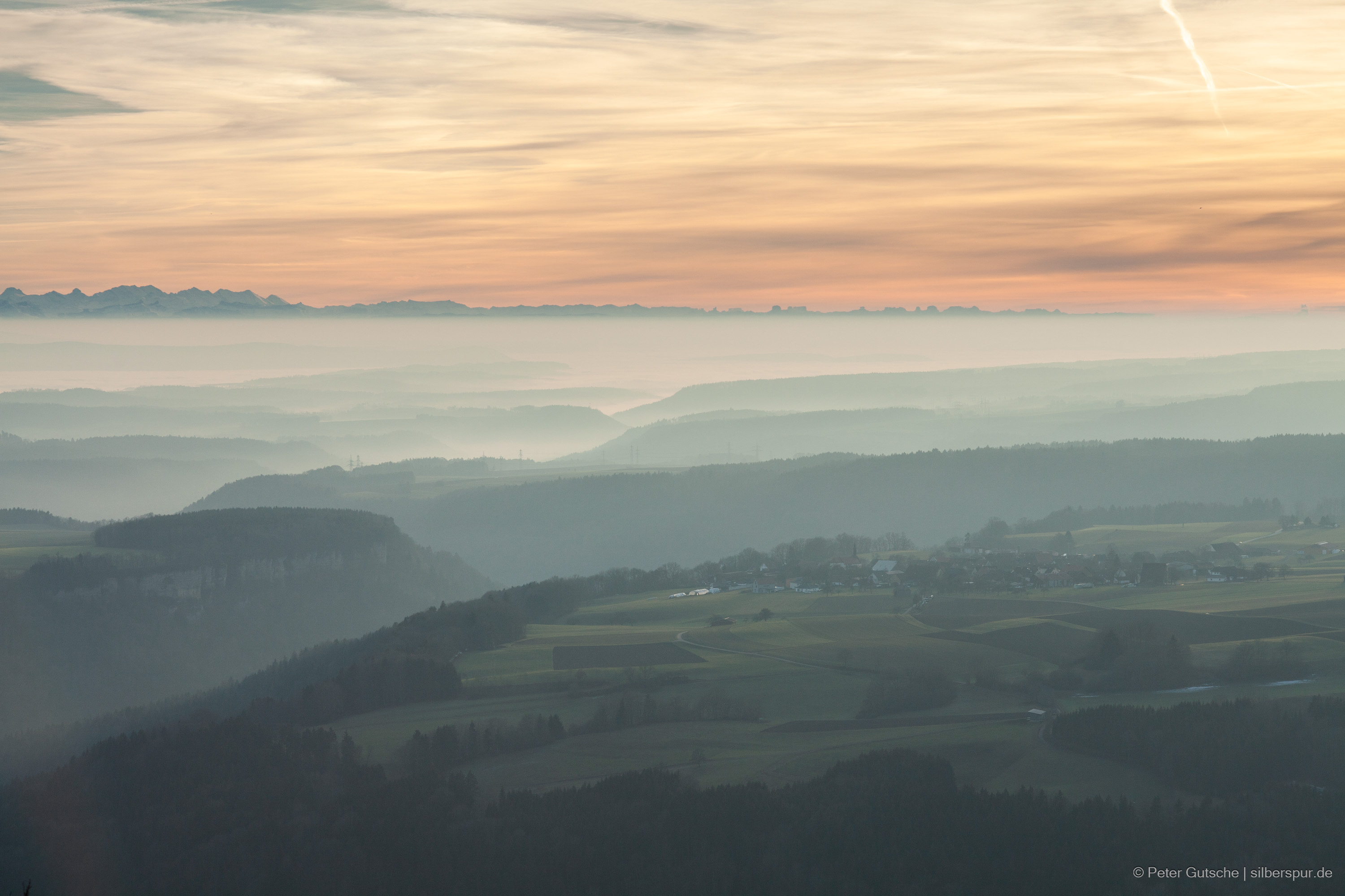 View from Buchberg in the Southern Black Forest towards the southwest against the afternoon sun. In the foreground, the lower hills of the Southern Black Forest are visible, with a village situated on the plateau. The low sun casts a warm light on the upper hilltops, while mist and fog linger in the valleys below. The plateau and hilly landscape are deeply carved by the valleys of the Wutach River and its tributaries. In the background, the majestic panorama of the Alps can be seen.