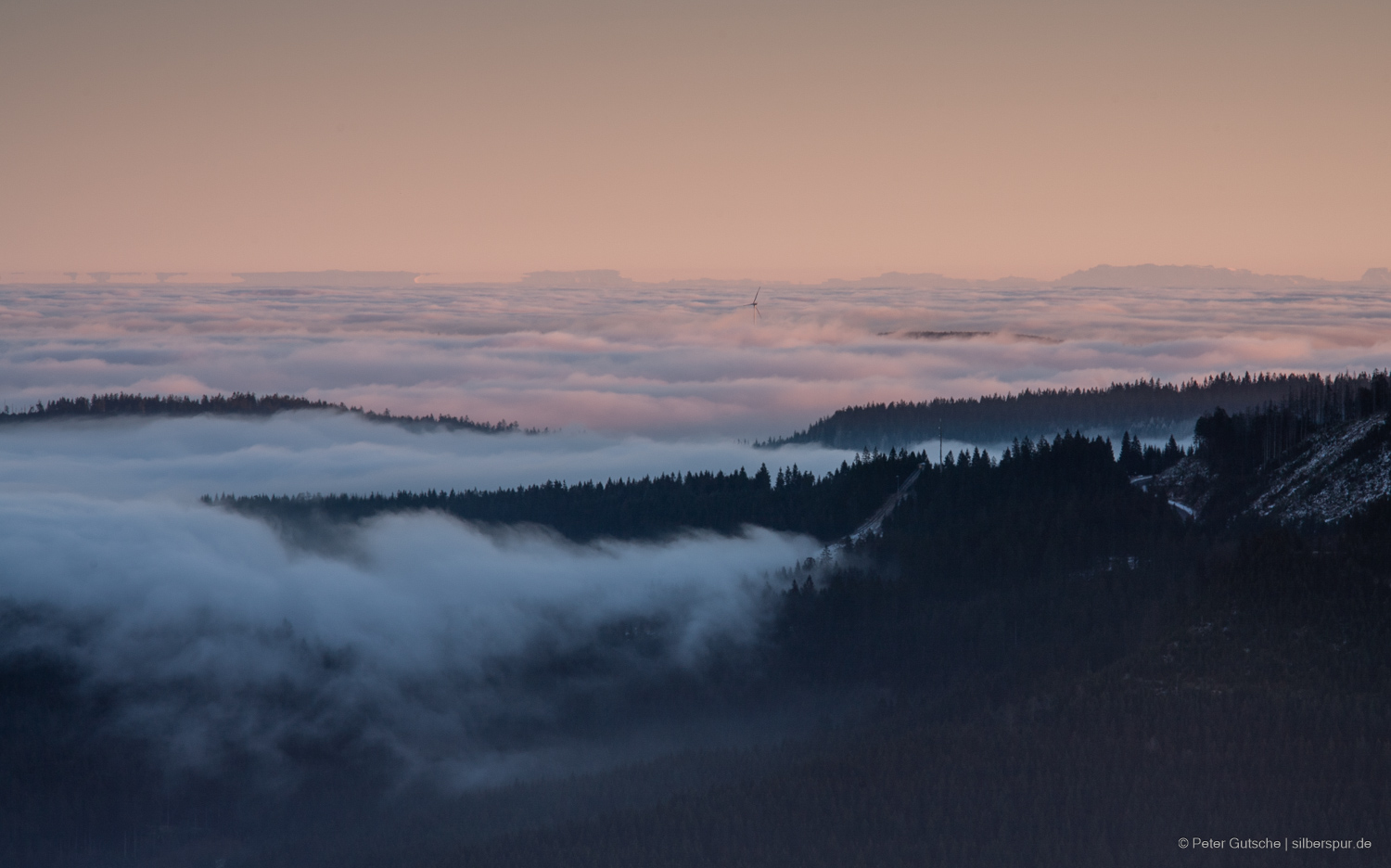 In the late afternoon sun, the view from a Black Forest peak extends over the neighboring slopes up to the distant Alps. In the valleys, fog lies due to a temperature inversion, appearing to spill over the neighboring mountain chain like a white cotton blanket. In the center of the image, a wind turbine rises above the sea of fog, while the silhouette of the Alps is distorted by optical phenomena known as mirages. Some parts of the Alps seem to float above the distorted peaks, suggesting a reflection.