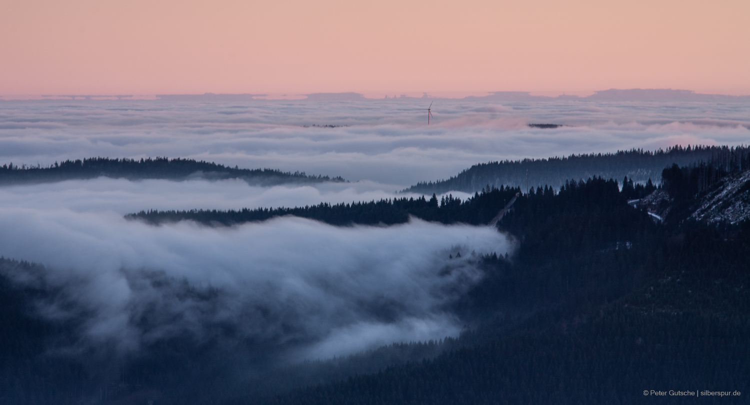 In the late afternoon sun, the view from a Black Forest peak extends over the neighboring slopes up to the distant Alps. In the valleys, fog lies due to a temperature inversion, appearing to spill over the neighboring mountain chain like a white cotton blanket. In the center of the image, a wind turbine rises above the sea of fog, while the silhouette of the Alps is distorted by optical phenomena known as mirages. Some parts of the Alps seem to float above the distorted peaks, suggesting a reflection. The silhouettes of the Alps have changed slightly compared to the previous picture.