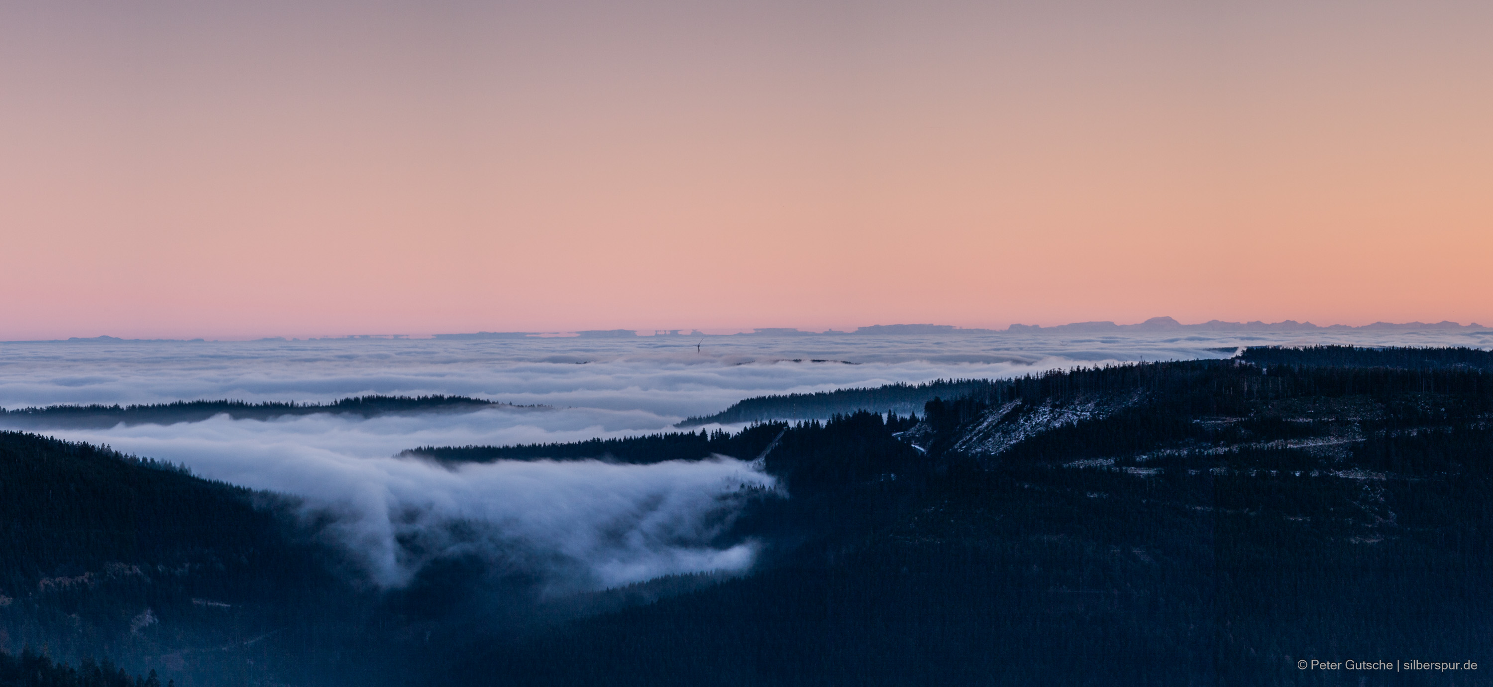 In the late afternoon sun, the view from a Black Forest peak extends over the neighboring slopes up to the distant Alps. In the valleys, fog lies due to a temperature inversion, appearing to spill over the neighboring mountain chain like a white cotton blanket. In the center of the image, a wind turbine rises above the sea of fog, while the silhouette of the Alps is distorted by optical phenomena known as mirages. Some parts of the Alps seem to float above the distorted peaks, suggesting a reflection. The silhouettes of the Alps have changed slightly compared to the previous picture. Compared to the previous images, this picture shows a broader section of the Alpine panorama.