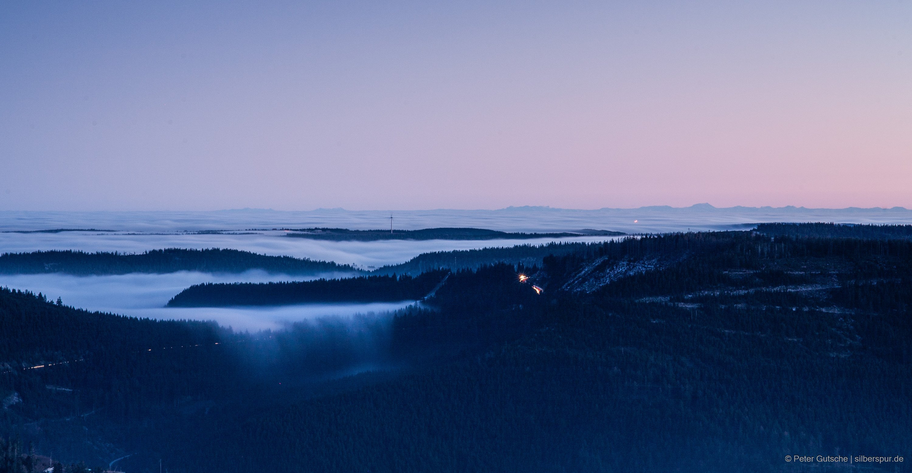 At dusk, the view from a Black Forest peak extends over the neighboring slopes up to the distant Alps. In the valleys, fog lies due to a temperature inversion, appearing to spill over the neighboring mountain chain like a white cotton blanket. In the center of the image, a wind turbine rises above the sea of fog. As the photo was taken later at dusk, the mirages of the distant Alps have vanished and the silhouettes of the Alps are undistorted.