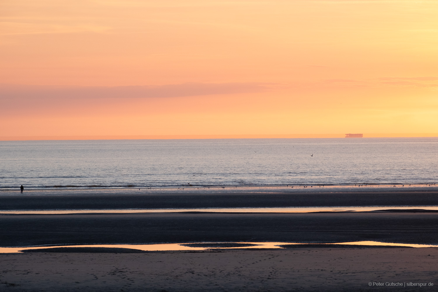 A coastal scene at low tide bathed in the warm glow of the setting sun. The beach is interspersed with puddles that reflect the vibrant red hues of the evening sky. In the distance, at the very left of the picture, a silhouette of a person stands at the shore, gazing out at the sea. On the horizon, at the very right of the picture, the outline of a container ship is visible, with its hull reflected, creating the illusion that the ship appears taller.
