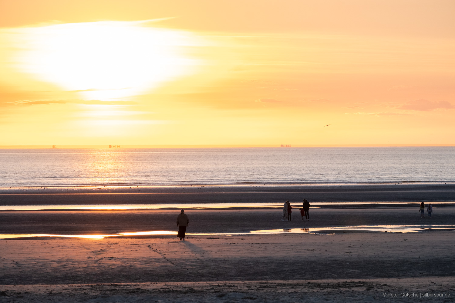 A coastal scene at low tide illuminated by the light of the low-hanging sun, which is visible in the frame. The beach is dotted with puddles that reflect the sunlight, creating a shimmering effect. Silhouettes of several people stand at the shore, gazing out at the sea. On the horizon, the outlines of three ships are visible, their shapes vertically mirrored by the phenomenon of mirage, enhancing the serene beauty of the scene.