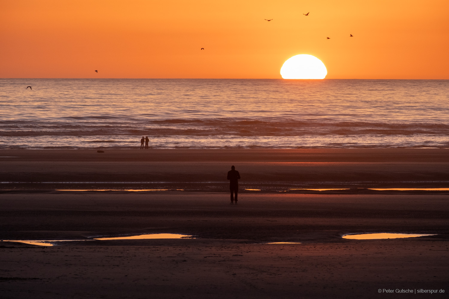 A coastal scene at low tide during sunset, captured with a strong telephoto lens, making the sun appear as a large, distorted ball, nearly half submerged on the horizon. The shape of the sun is significantly compressed vertically, resembling overlapping layers of varying widths. The beach is interspersed with puddles that reflect the warm light of the setting sun. Silhouettes of several people stand at the shore, gazing out at the sea, adding a sense of tranquility to the dramatic landscape. There are also the silhouettes of some birds flying in front of the bright sky.