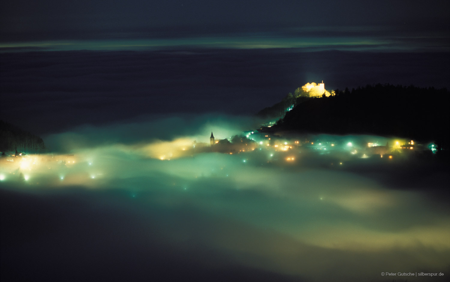 Nighttime view from a Black Forest mountain overlooking a foggy mountain saddle, where a small, illuminated village nestles under a sky of inversion weather conditions. Thick fog blankets the lower Rhine Valley and the surrounding Black Forest valleys, reaching up to the village’s edge. Soft, artificial lights from the village homes create a warm glow that partly penetrates the mist, casting a surreal, dreamlike effect. Higher up, an illuminated castle crowns a nearby mountain peak, standing out above the fog-covered landscape.