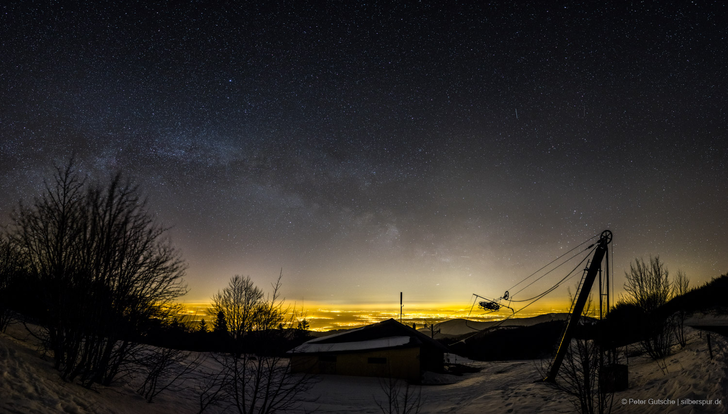 The framework of a ski lift at night on a mountain summit. In the background, the brightly illuminated plain, above it the starry sky with the band of the Milky Way.