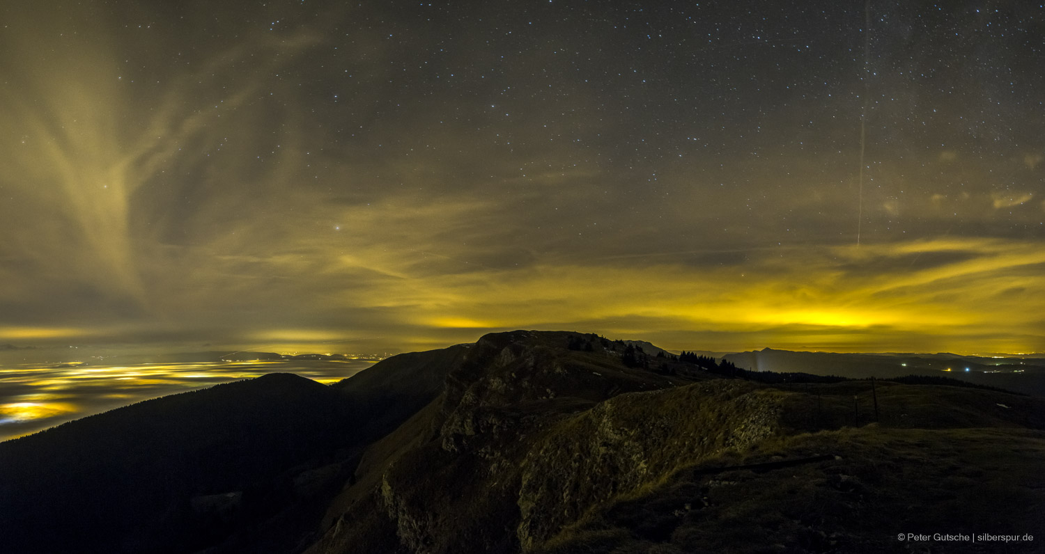 A dark mountain ridge at night, behind which parts of the more distant landscape can be seen on the left and right. To the left, a sea of fog, beneath which yellowish city lights shine through. Stars are visible in the sky, but also large cirrus clouds reflecting the light of the cities.