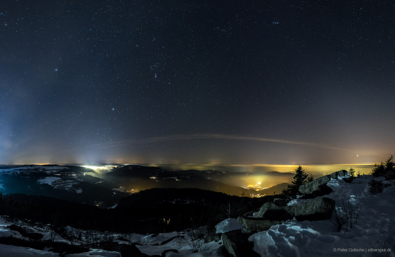 View from a mountain peak on a winter night into the neighboring valleys and the distant Rhine plain. In the valleys, the lights from villages and towns are shining, partially under the fog. In the sky, which makes up about two-thirds of the image, many stars are visible. On the left side of the image, the blue lights from ski lifts shine particularly brightly.