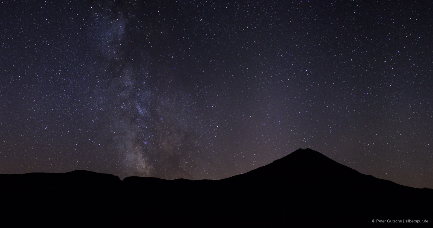 A dark mountain silhouette at night with the starry sky above. 
                The bright band of the Milky Way is clearly seen in the center of the image 
                 as it rises perpendicular into the sky.