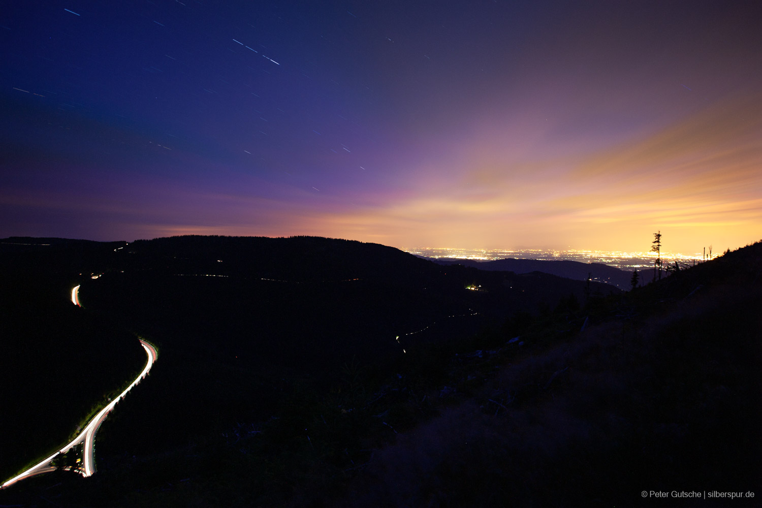 View from a higher mountain in the northern Black Forest overlooking the slopes traversed by the Black Forest High Road. The lights of individual cars traveling in the dark create light trails along the shadowed mountainside, as the photo was taken with a multi-minute exposure. In the background, the Rhine Plain is brightly illuminated by the artificial lights of the cities. In the partly cloudy sky, a few star trails are visible.