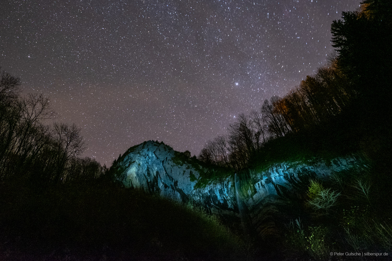 A limestone rock in the French Jura is illuminated at night by a headlamp, highlighting the visible sediment layers and folds. Above, the sky is filled with stars.