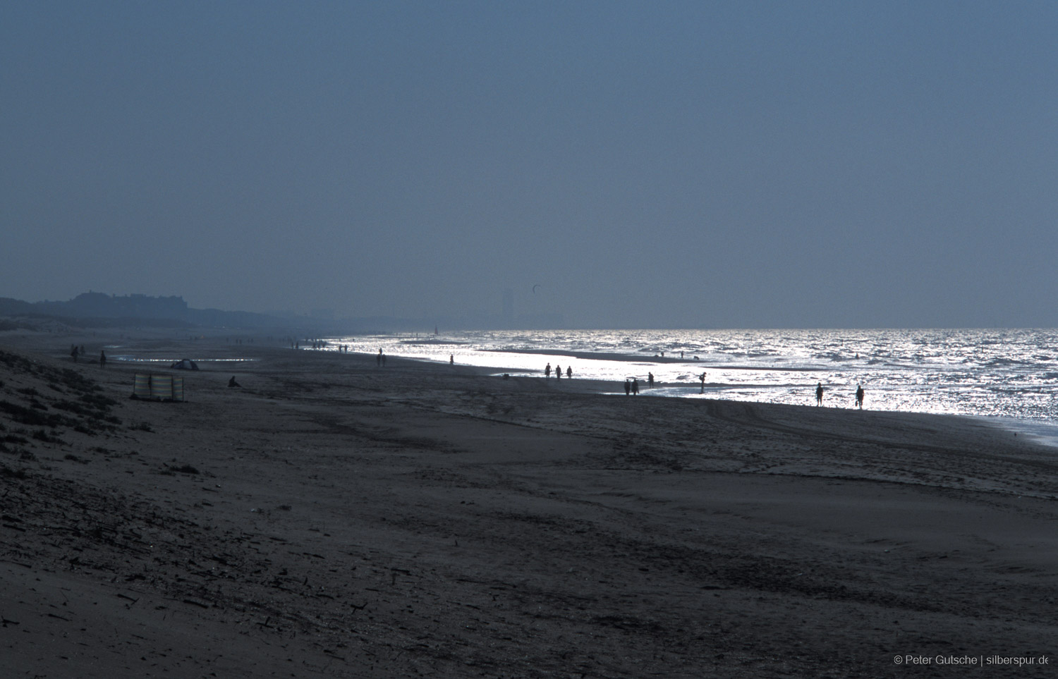 A beach photographed against the sunlight with the dazzling, shimmering sea in the background. In front of the silvery shine, the silhouettes of individual walkers stand out, giving the image a touch of mystery.