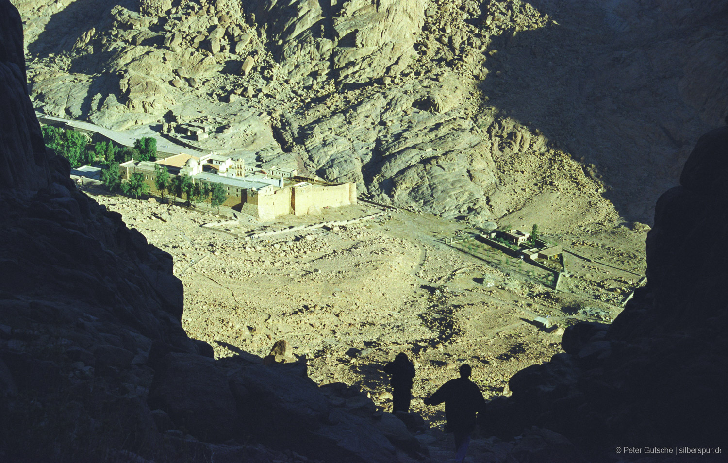 A mountainous desert landscape bathed in yellow light. In the foreground, the scene is framed by shaded mountain slopes to the left and right. In the background, the opposite cliffs are brightly lit by the hot sun, with part of St. Catherine's Monastery nestled in the valley below. In front of the viewer, the silhouettes of two hikers appear, also making their way down the descent.