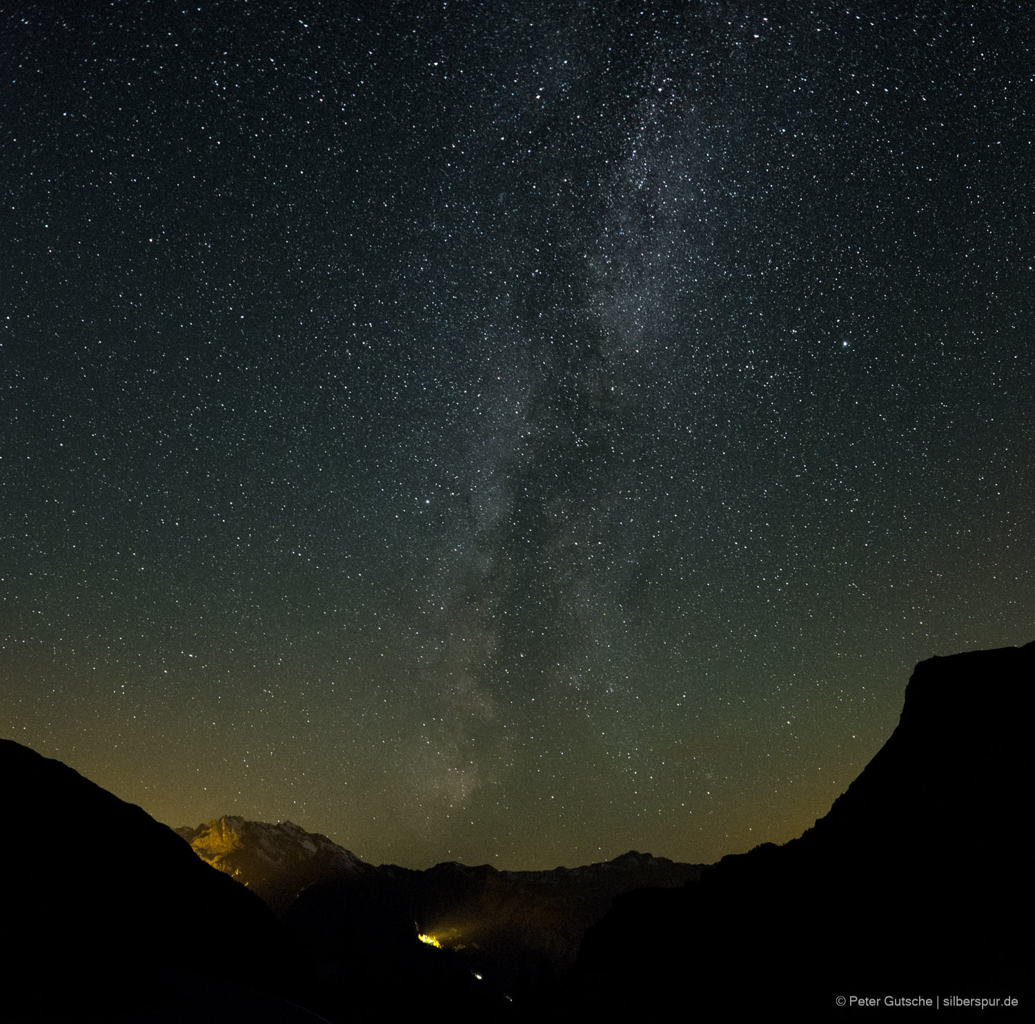 Night photo taken in the Alps with the Milky Way clearly visible in the sky. The mountain slopes in the foreground are partly illuminated by a construction site.