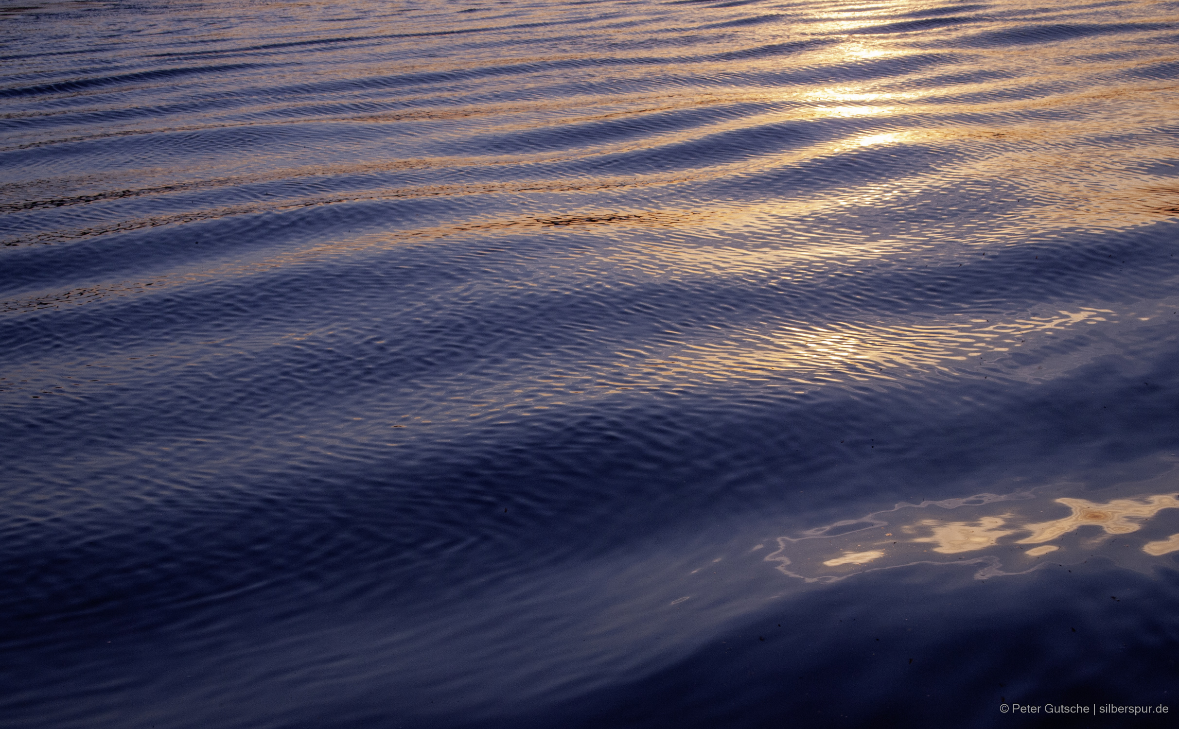 Eine von Wellen verformte Wasseroberfläche gegen die untergehende Sonne fotografiert. Der Bildausschnitt zeigt jedoch lediglich die Wasseroberfläche. Da es sich um leichten Wellengang handelt, wirkt das Bild eher ruhig.