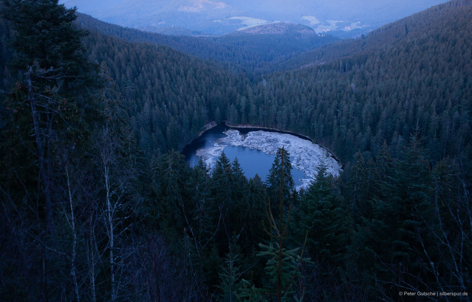 From a mountain slope, you look down to the Huzenbacher See with its nearly cirque shape. The lake is located in a depression and surrounded by a deep forest. The surface of the lake is partly covered by areas of moor. As the photo was made in winter, these areas are partly covered by hoarfrost shing brightly.