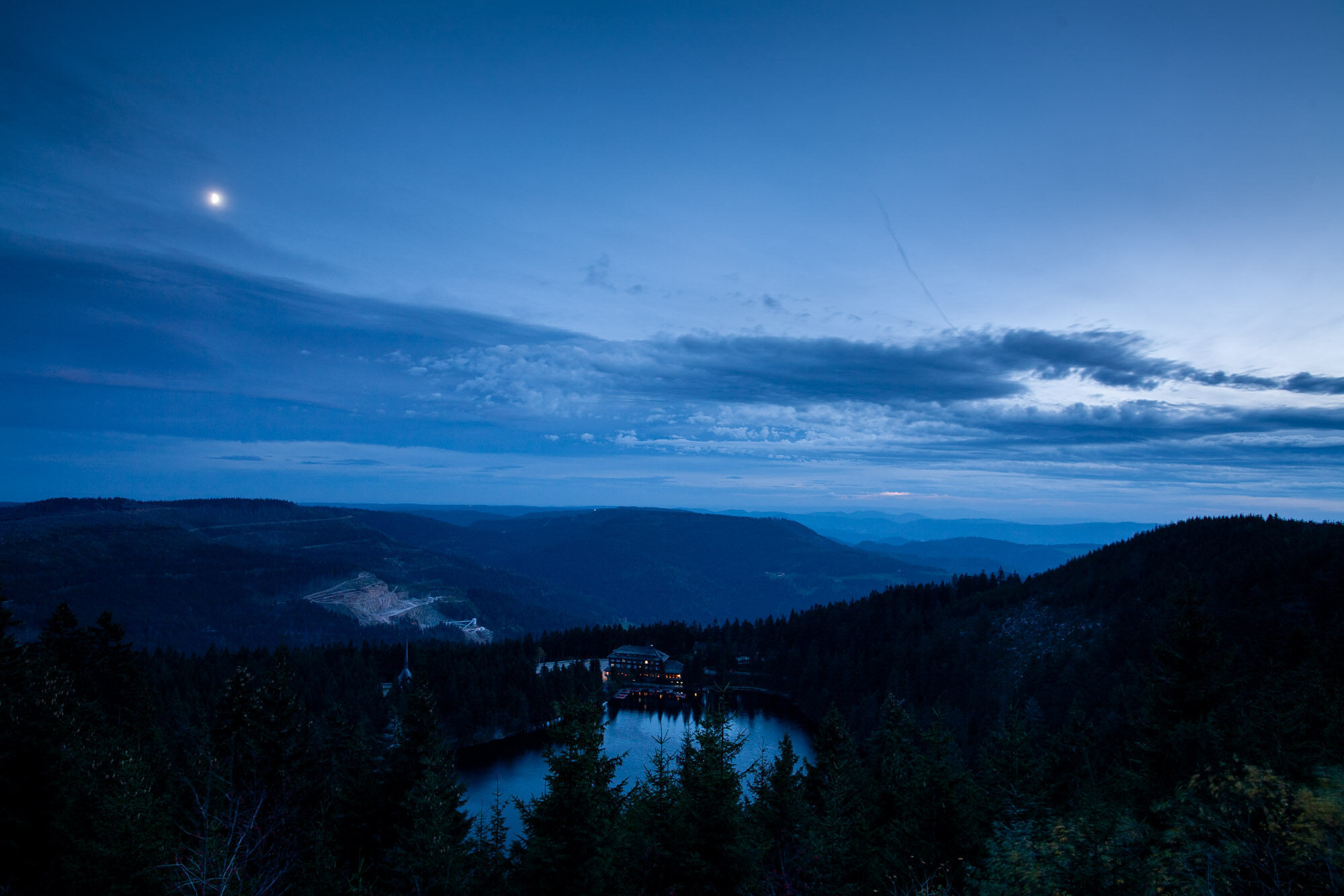 A cirque lake at dusk seen brom above. There’s a hotel at the shore of the lake with illuminated windows. The picture is in dark blue with dark clouds developing at the sky. In a clear area of the sky, the moon shows up.