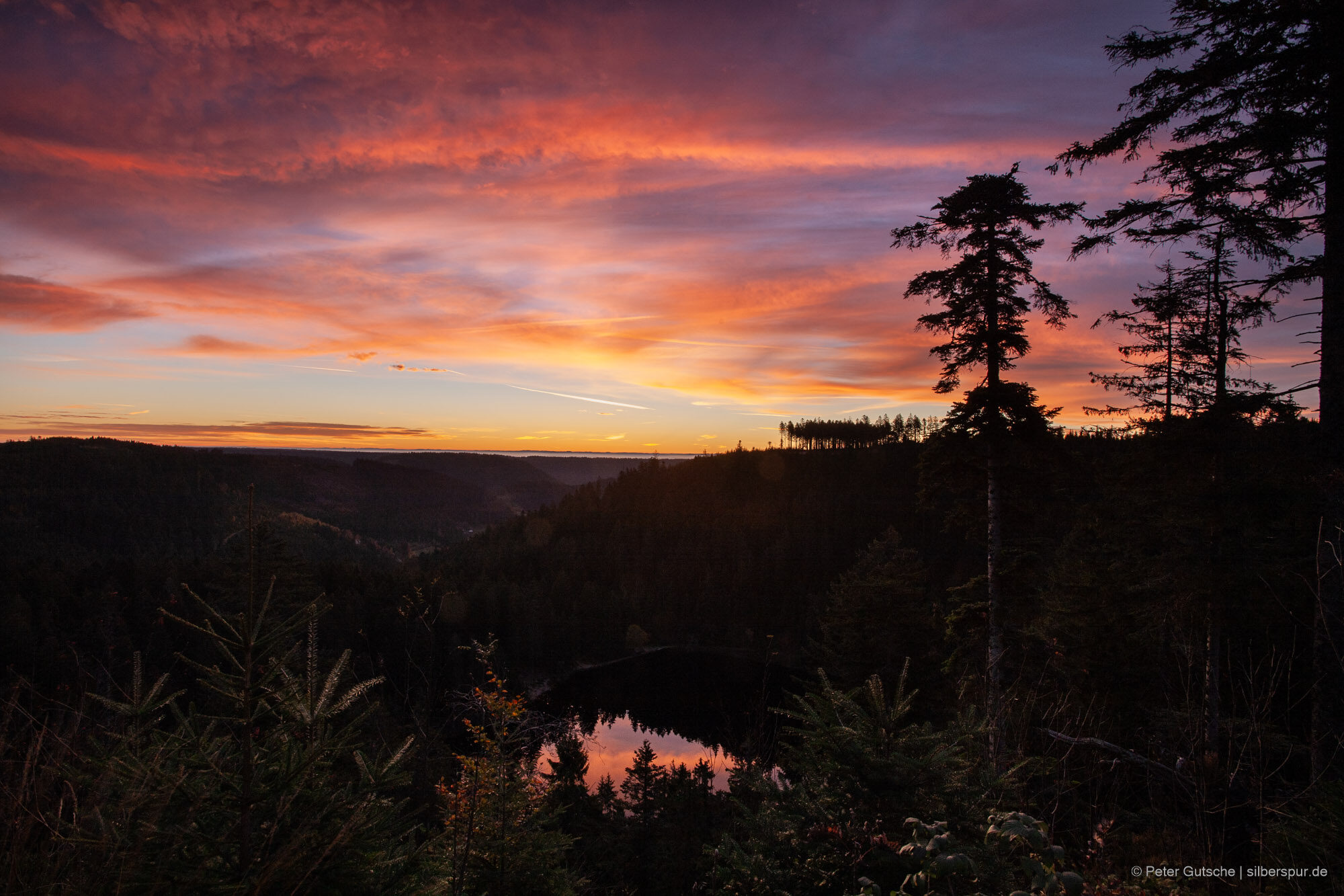 A photo taken at dawn. Dark mountain ridges under a red sky. In the dark below, a tiny lake reflects the light of the sky.