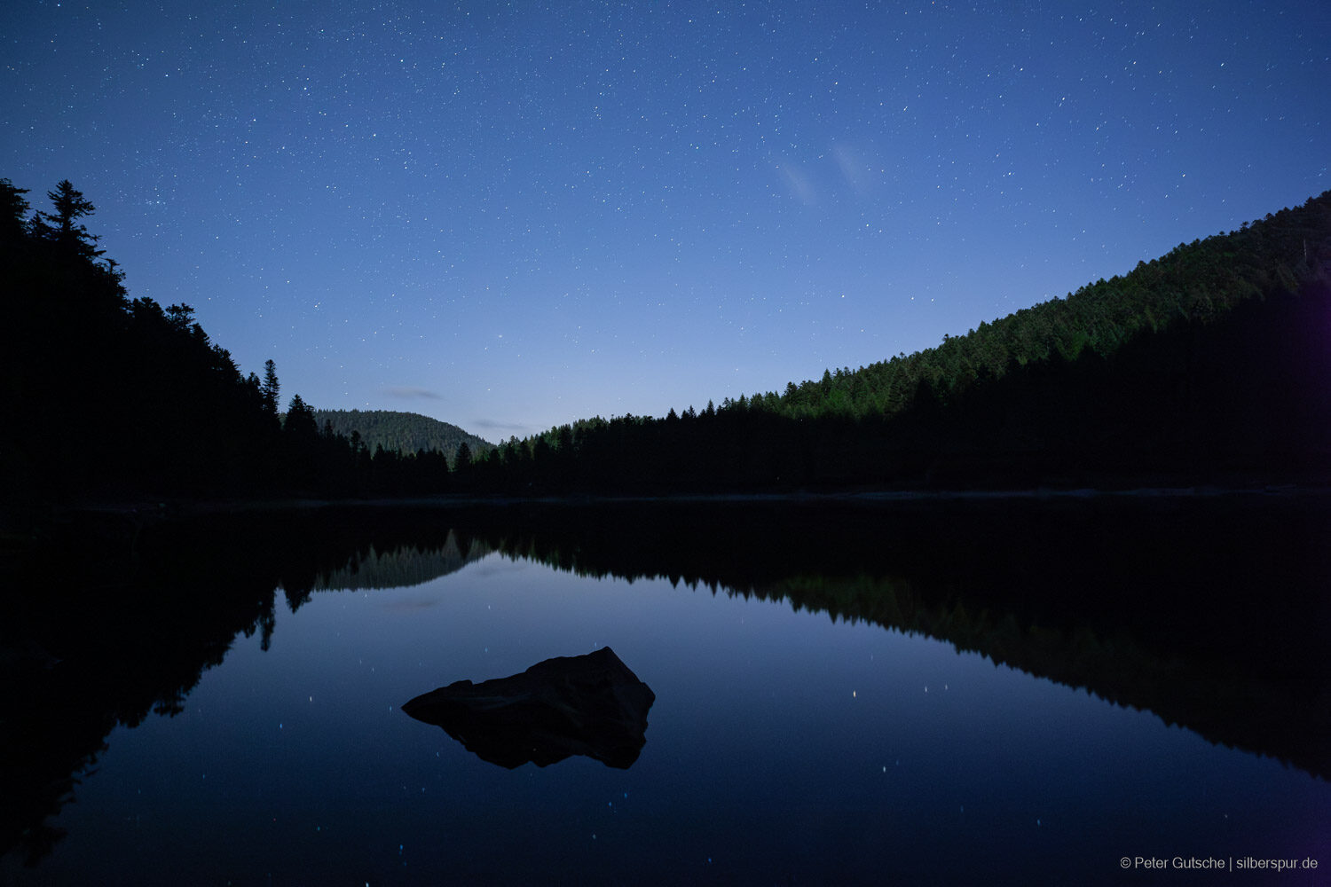 A lake at night with the starry sky reflected on the water surface. A rock protrudes from the water which is a black shape.
